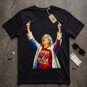 May include: A black short-sleeved t-shirt featuring a graphic of a person with blue hair, glasses, and a light jacket, raising both middle fingers. The person wears a red shirt with the text PRIMUS NO KINGS. A brown paper tag is attached to the collar.