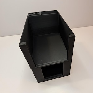 May include: A matte black 3D printed storage organiser featuring a large rectangular main compartment, a smaller rectangular section, and two circular slots at the top. The structure has a hollow base opening and a textured surface finish.