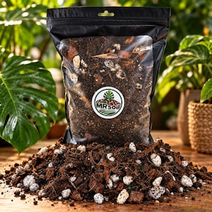 May include: A black plastic bag filled with chunky tropical potting soil mix, featuring a circular white label with the text MR soil and a green leaf graphic. The bag sits behind a pile of the same dark brown soil, wood chips, and white perlite.