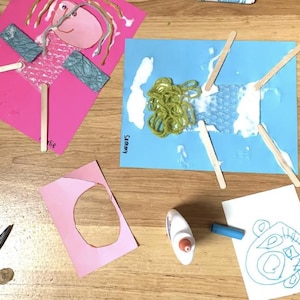 May include: Two children's art projects on pink and blue paper featuring popsicle stick limbs, yarn hair, and bubble wrap textures. One project is labelled 'Sammy' and the other 'Ellie'. Includes a glue bottle, blue crayon, scissors, and a drawing.