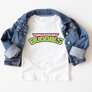 May include: White t-shirt with the text "TEENAGE MUTANT NINJA BUDDIES" in green and red, laid on top of a blue denim jacket. The shirt's design is reminiscent of a popular cartoon.