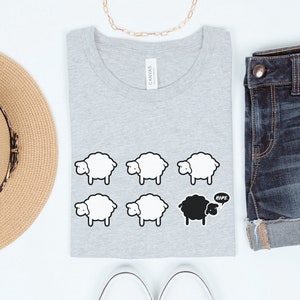 May include: A gray t-shirt with a black sheep and five white sheep printed on it. The black sheep has a speech bubble that says "Nope". The t-shirt is folded and laid flat on a white surface. There is a straw hat, a pair of white sneakers, and a pair of blue denim shorts in the image.