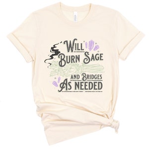 May include: A beige t-shirt with the text "Will Burn Sage and Bridges As Needed" in black and purple lettering. The text is surrounded by a stylized image of smoke and crystals.