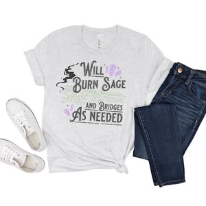 May include: A heather grey t-shirt with a graphic design that says "Will Burn Sage and Bridges As Needed". The graphic includes a black silhouette of a witch's head, a bundle of sage, and purple crystals.