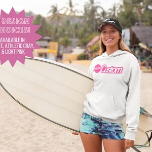 May include: A white hoodie with a pink circle and the word "Custom" in pink lettering. The hoodie is being worn by a person holding a surfboard. The text "3 DESIGN CHOICES! AVAILABLE IN: WHITE, ATHLETIC GRAY, & LIGHT PINK" is in a pink starburst shape.