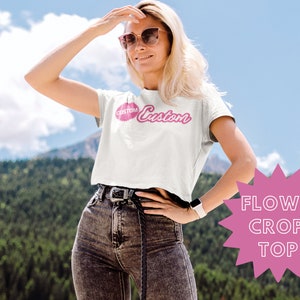 May include: A white crop top with a pink "Custom" graphic. The top is flowy and has short sleeves. The model is wearing the top with black jeans and sunglasses.