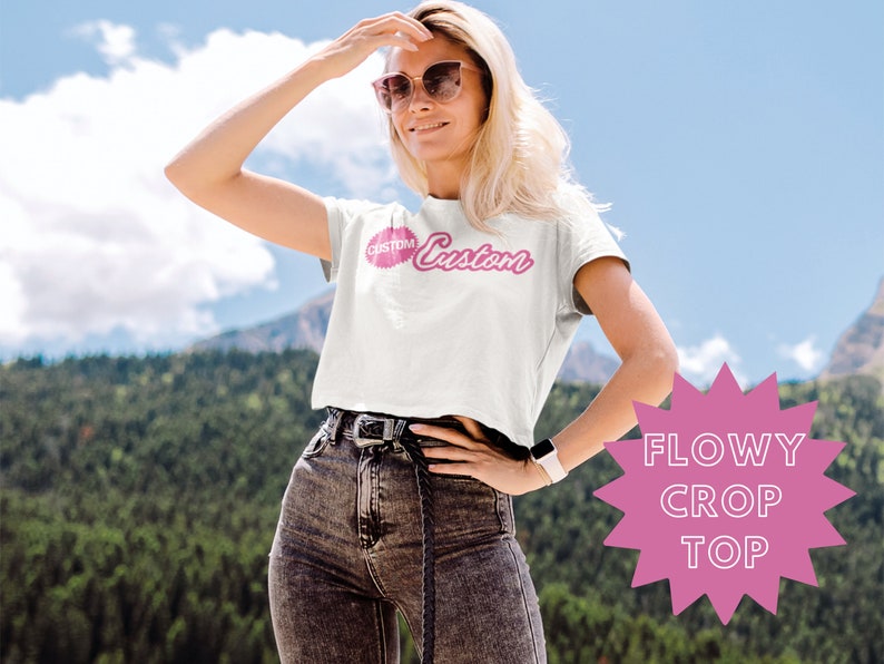 May include: A white crop top with a pink "Custom" graphic. The top is flowy and has short sleeves. The model is wearing the top with black jeans and sunglasses.