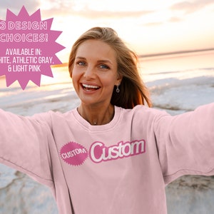 May include: A woman wearing a light pink sweatshirt with the word "Custom" printed on it. The sweatshirt is available in white, athletic gray, and light pink.