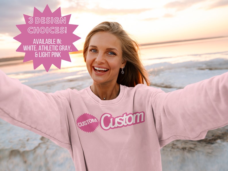 May include: A woman wearing a light pink sweatshirt with the word "Custom" printed on it. The sweatshirt is available in white, athletic gray, and light pink.