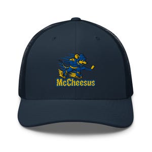 Op de afbeelding: Een marineblauwe truckerpet met een geborduurde afbeelding van een gele en blauwe muis in ijshockeytenue met schaatsen en een ijshockeystick. Onder de afbeelding is de tekst McCheesus in het geel met blauwe omlijning geborduurd.