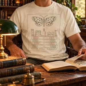 May include: An off-white short-sleeve t-shirt featuring a black line-art graphic of a moth above a stack of books, a candle, and spectacles. The text "PLATE LIX Bibliotheca The Reading Room" is printed at the bottom of the rectangular illustration.