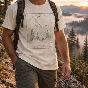 Może przedstawiać: Kremowy t-shirt z krótkim rękawem z czarną grafiką liniową przedstawiającą półksiężyc, gwiazdy i drzewa iglaste w prostokątnej ramce. Pod ilustracją znajduje się napis: PLATE CXIII - Luna Montium - The Mountain Moon.