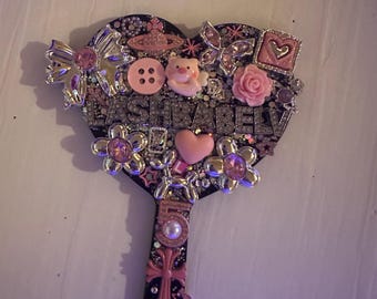 Pode incluir: Um espelho de mão em forma de coração decorado com adornos cor-de-rosa e prateados, incluindo um laço, um porquinho, uma rosa, flores, uma cruz e o texto LASHBABELV. A superfície está coberta de purpurina e vários berloques, incluindo um número cinco.