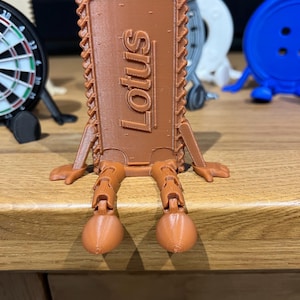 May include: A brown 3D printed figure shaped like a rectangular Lotus biscuit with a serrated border. The figure features articulated arms and legs with rounded feet, and the word Lotus is embossed vertically in the centre of the biscuit body.