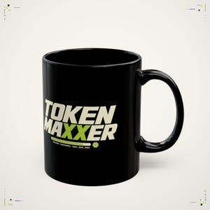 May include: A black ceramic mug featuring the white and lime green text TOKEN MAXXER. Below the text is a lime green progress bar and the small white text TOKENS CONSUMED: 999,999,999 next to a small white microchip icon on the side of the mug.
