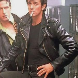May include: A black leather motorcycle jacket with silver metal zippers and snaps, worn over a plain black t-shirt. The jacket features a wide collar, lapels, and long sleeves. The person is wearing black pants with a black belt and has dark hair.