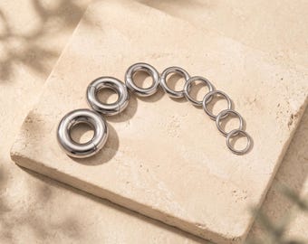 Puede incluir: Un conjunto de ocho anillos metálicos circulares de tono plateado dispuestos en una línea curva de mayor a menor. Cada anillo presenta una superficie lisa y pulida con un diseño sin costuras. El nombre de la marca LUJAIN es visible en la esquina inferior derecha.