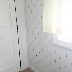 May include: A light gray wall covered in a repeating pattern of small pink rosebuds with green stems. The wall features a white door on the left, a white window frame with a sheer white curtain on the right, and a white electrical outlet.