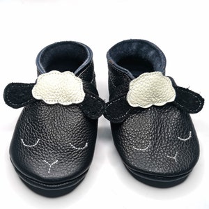 Può includere: Un paio di scarpine per neonati in pelle nera con un design a forma di pecora. Le scarpe hanno una toppa di lana bianca sulla parte superiore e orecchie in feltro nero.