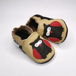 Puede incluir: Un par de zapatos de bebé de cuero beige con un diseño de búho. Los búhos son negros con alas rojas y ojos blancos. Los zapatos tienen un forro de cuero marrón y una suela negra. El nombre de la marca "Ebooba" está impreso en el lateral de los zapatos.