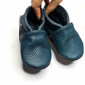 May include: A pair of teal blue leather baby shoes with a white stitch detail and a black sole. The shoes have a soft, flexible design and are perfect for little feet.