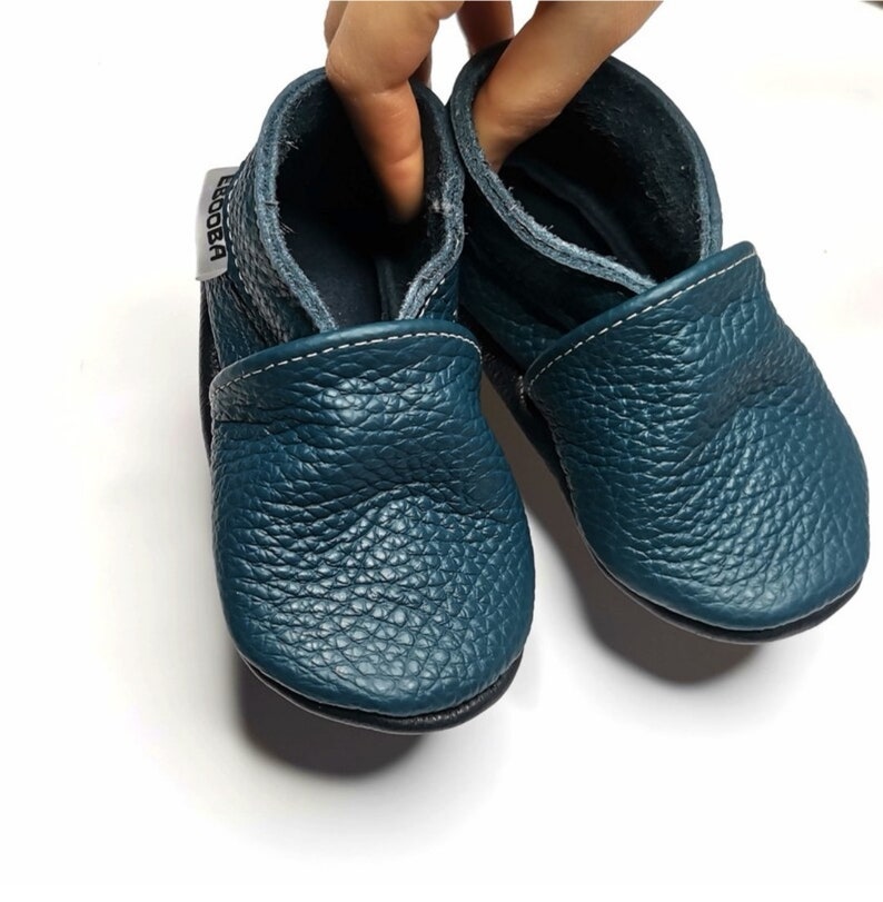 May include: A pair of teal blue leather baby shoes with a white stitch detail and a black sole. The shoes have a soft, flexible design and are perfect for little feet.