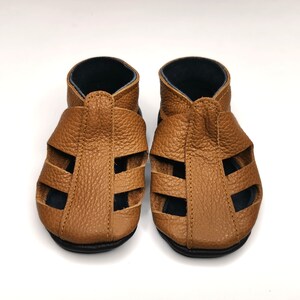 May include: A pair of brown leather sandals with a criss-cross strap design. The sandals have a black lining and a closed toe.