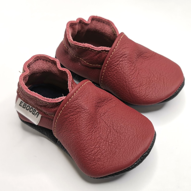 May include: A pair of burgundy leather baby shoes with a black sole and a white label that says "Ebooba".