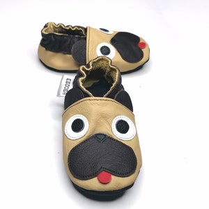 May include: A pair of tan and brown baby shoes with a pug dog face design. The shoes have black noses, ears, and eye details, with white eye surrounds and a red tongue. The shoes are made of leather.