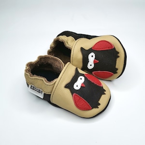 May include: A pair of tan leather baby shoes with an owl design. The owl is black with red wings and white eyes. The shoes have a brown leather lining and the brand name "Ebooba" is printed on the side.