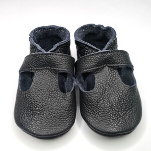 May include: A pair of black leather baby shoes with a strap across the top and a navy blue lining.
