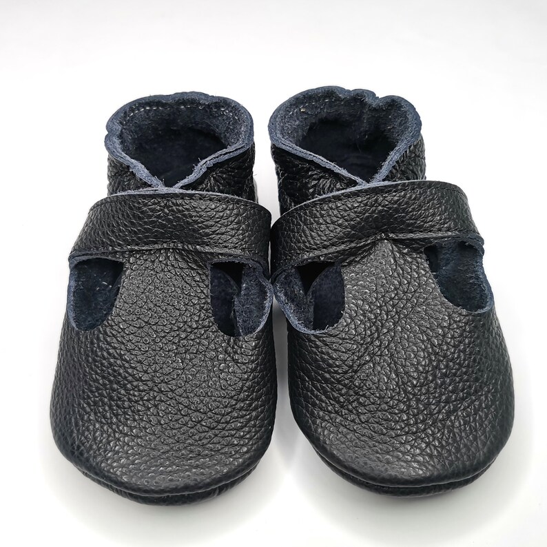 May include: A pair of black leather baby shoes with a strap across the top and a navy blue lining.