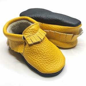 Peut inclure: Une paire de mocassins pour bébé en cuir jaune avec des détails en frange. Les mocassins ont une semelle noire et sont faits de cuir souple et doux.