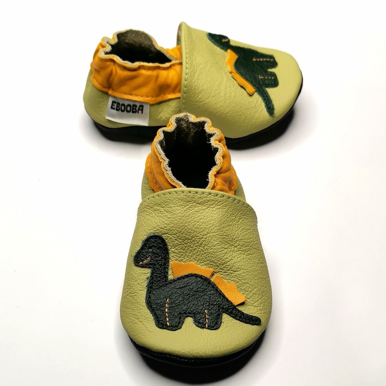 May include: A pair of green leather baby shoes with a dinosaur design. The shoes have a yellow lining and the word "Ebooba" is printed on the side.