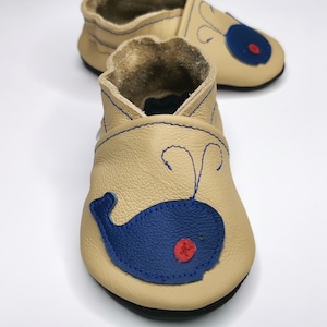 Może przedstawiać: Beżowe skórzane buciki dla niemowląt z wzorem niebieskiego wieloryba i czerwonym guzikiem w roli oka. Wieloryb ma niebieski kontur i biały brzuch.