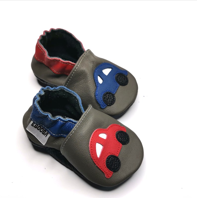 May include: A pair of gray leather baby shoes with a blue and red car design. The shoes have a soft, flexible sole and are perfect for little feet.