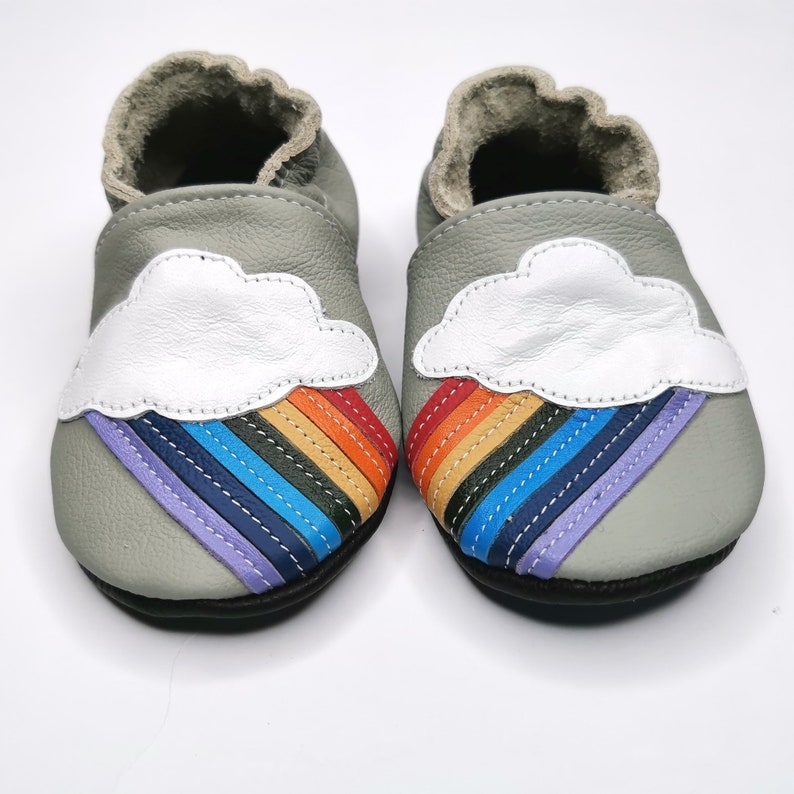 May include: Gray leather baby shoes with a white cloud and rainbow design. The rainbow is made of colorful stitching.
