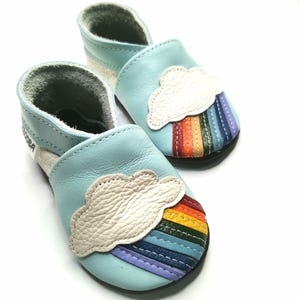 May include: A pair of light blue leather baby shoes featuring a white cloud and rainbow design. The shoes are soft and flexible, ideal for little feet.