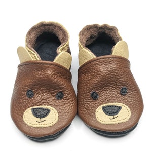 May include: A pair of brown leather baby shoes with a bear face design. The shoes have a tan colored trim around the edges and a black sole.
