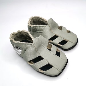 May include: A pair of light gray leather baby sandals with an open toe and a strap across the foot. The sandals have a black sole and a label that says "Ebooba".