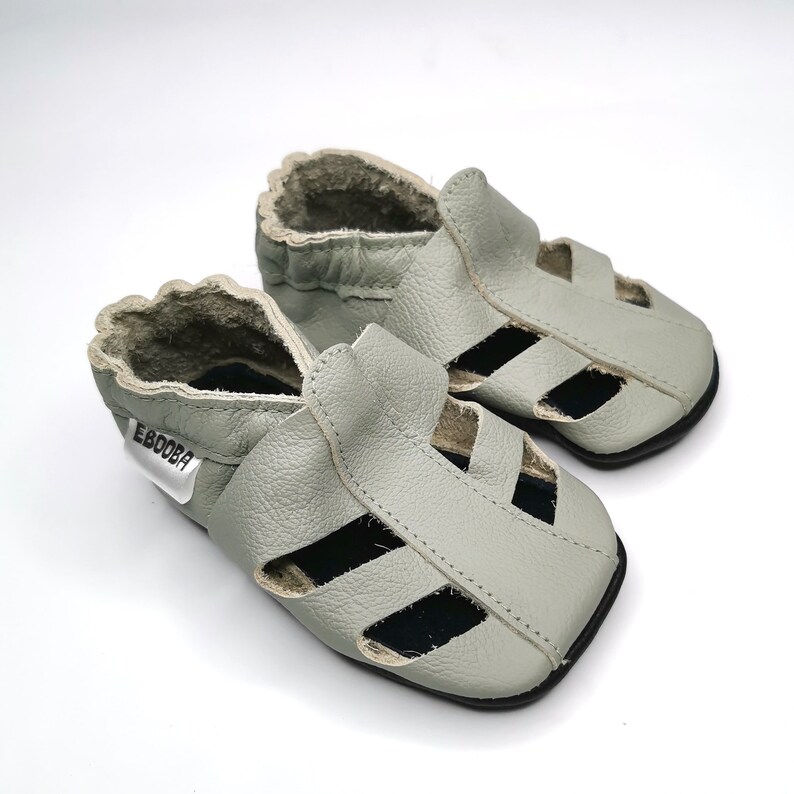 May include: A pair of light gray leather baby sandals with an open toe and a strap across the foot. The sandals have a black sole and a label that says "Ebooba".