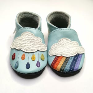 以下が含まれることがあります： 片方の靴に白い雲と雨滴、もう片方の靴に虹が描かれた、薄い水色のベビーシューズのペアです。