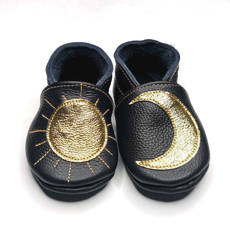 May include: A pair of black leather baby shoes with gold embroidered sun and moon designs.