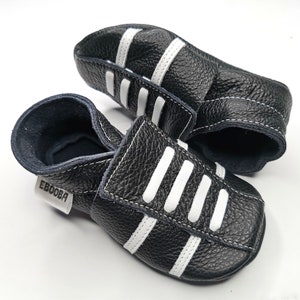 May include: A pair of black leather baby shoes with white stripes. The shoes have a velcro strap closure and the brand name "Ebooba" is printed on the side.