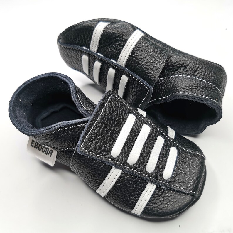 May include: A pair of black leather baby shoes with white stripes. The shoes have a velcro strap closure and the brand name "Ebooba" is printed on the side.