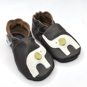 K&ouml;nnte beinhalten: Ein Paar braune Lederschuhe f&uuml;r Babys mit einem wei&szlig;en Elefanten-Design und einem gelben Knopf an jedem Schuh. Die Schuhe haben eine weiche, flexible Sohle und sind perfekt zum Krabbeln und Laufen.