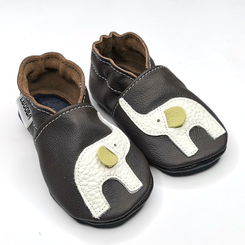 K&ouml;nnte beinhalten: Ein Paar braune Lederschuhe f&uuml;r Babys mit einem wei&szlig;en Elefanten-Design und einem gelben Knopf an jedem Schuh. Die Schuhe haben eine weiche, flexible Sohle und sind perfekt zum Krabbeln und Laufen.