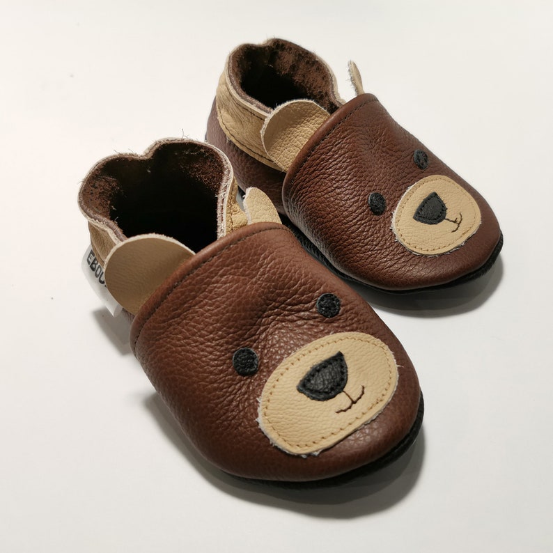 May include: A pair of brown leather baby shoes with a bear face design. The shoes have a soft, suede lining and a flexible sole.