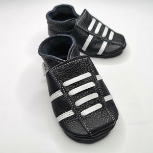 May include: Black leather baby shoes with white stripes. The shoes have a soft, flexible sole and are perfect for crawling and walking.