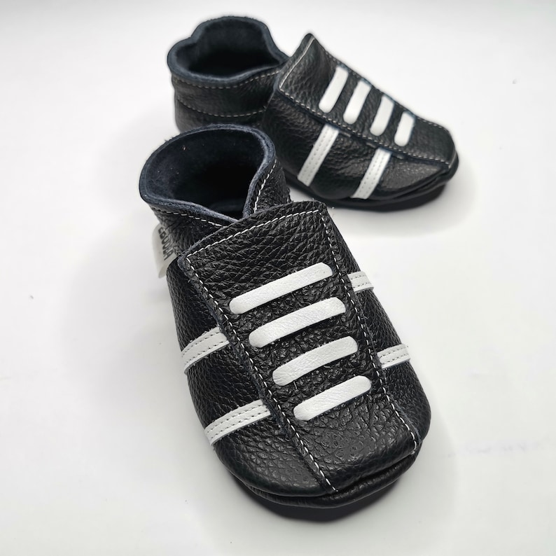 May include: Black leather baby shoes with white stripes. The shoes have a soft, flexible sole and are perfect for crawling and walking.